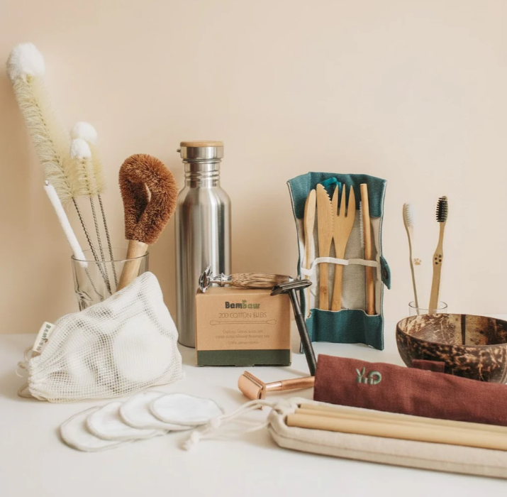 Collection of sustainable living tools including bamboo toothbrushes, reusable stainless steel water bottles, and cleaning brushes on a natural countertop.