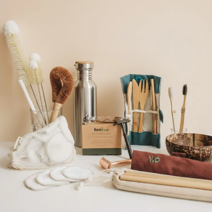 Collection of sustainable living tools including bamboo toothbrushes, reusable stainless steel water bottles, and cleaning brushes on a natural countertop.