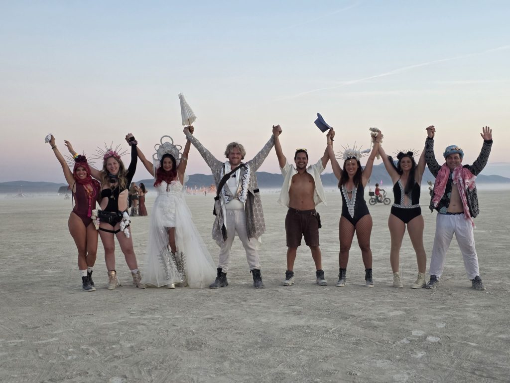 burning man sustainability image of people holding hands on the playa at burning man. All individuals have fun, creative costumes on during sunset. 