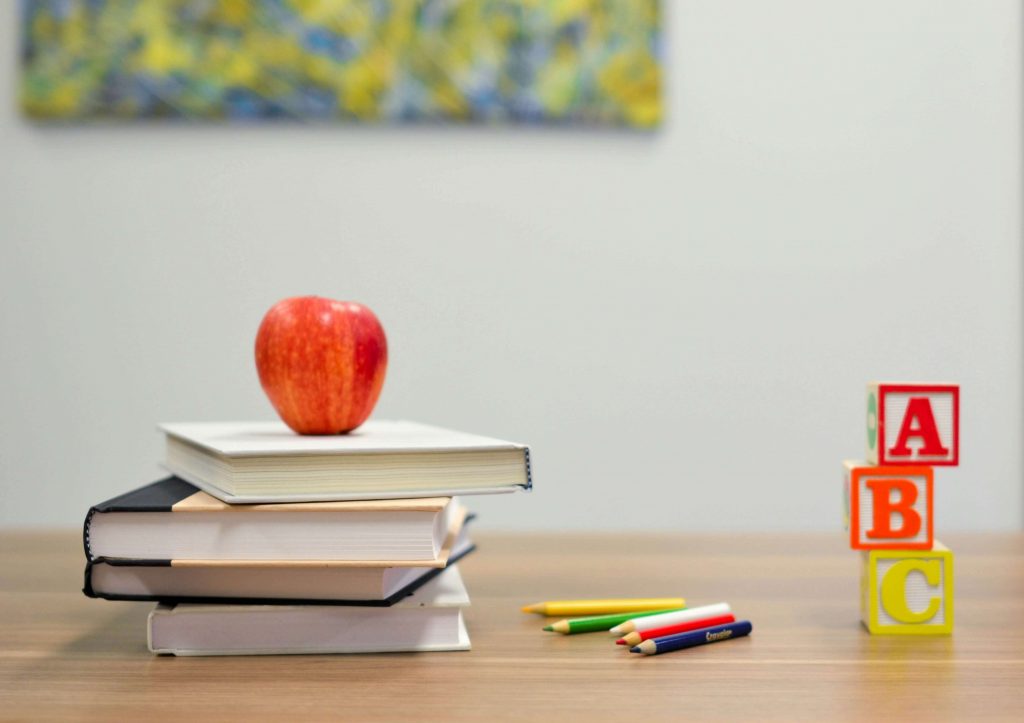 Stack of books with an apple on top and wooden ABC blocks on a classroom desk.