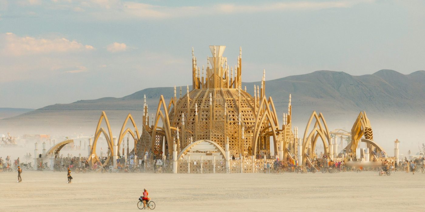 Is Burning Man sustainable? showcased with an image of a temple built in the middle of the desert and afterwards is taken down with the burning man "leave no trace" principles. Image of a wooden temple in the middle of Black Rock City.