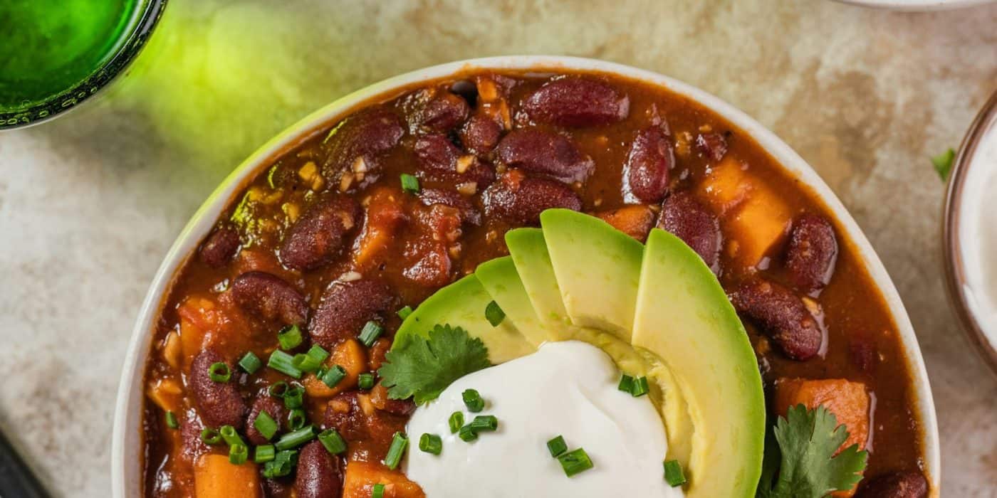 Image of best vegan chili plant-based with beans, veggies, avocado, and vegan sour cream on a table.