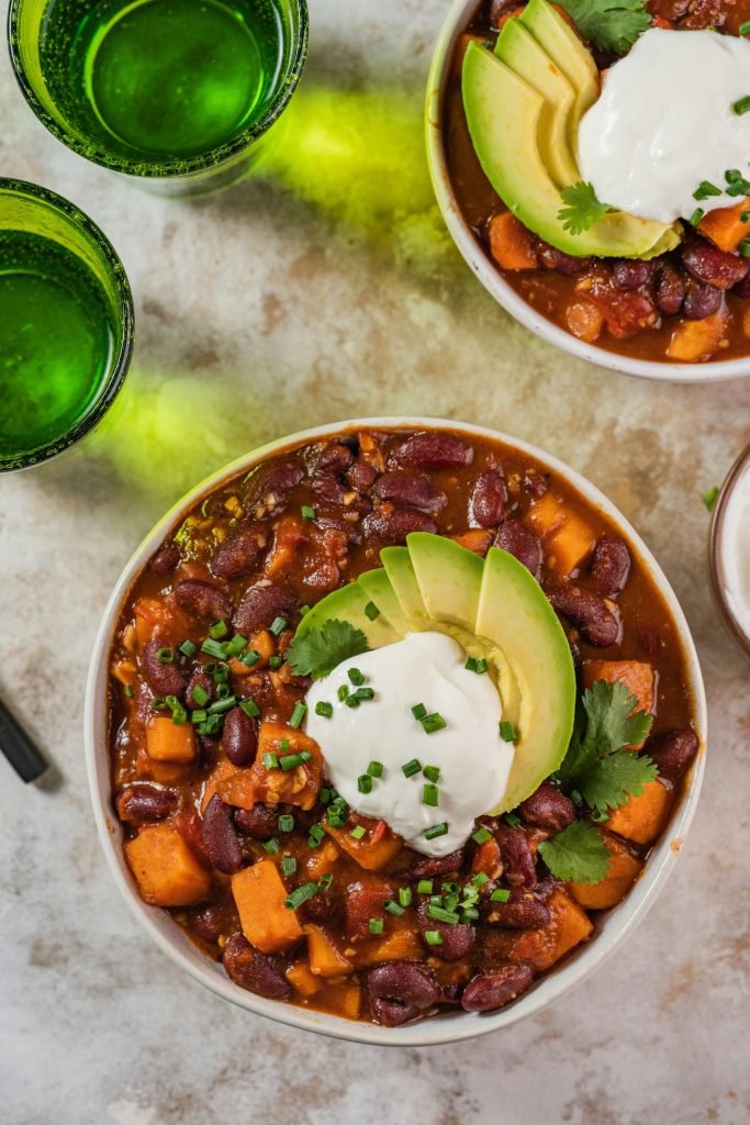 Image of best vegan chili plant-based with beans, veggies, avocado, and vegan sour cream on a table.